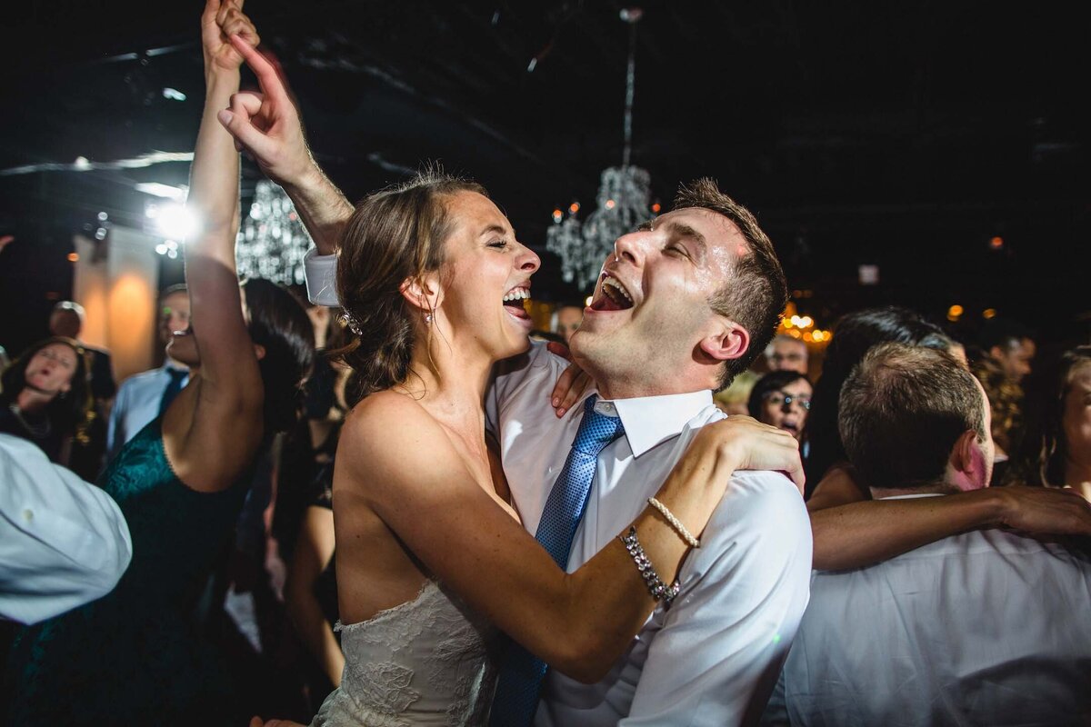 A newlywed couple shares a kiss after a Jewish wedding at the Boathouse at Confluence Park surrounded by guests holding sparklers at night, creating a magical and festive atmosphere. The bride's curly hair and the groom's beard are softly illuminated by the warm light of the sparklers.