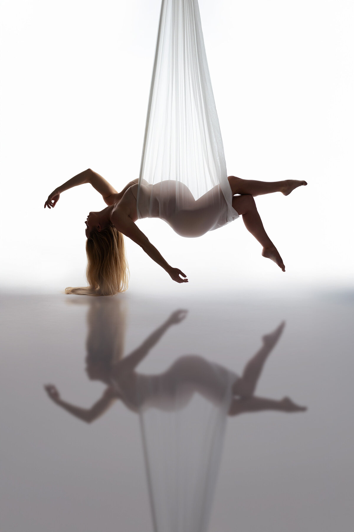 An elegant woman floats effortlessly while suspended in sheer white fabric, her body gently curved and beautifully reflected on the smooth surface below. This ethereal maternity photograph showcases the artistry of DFW maternity photography.