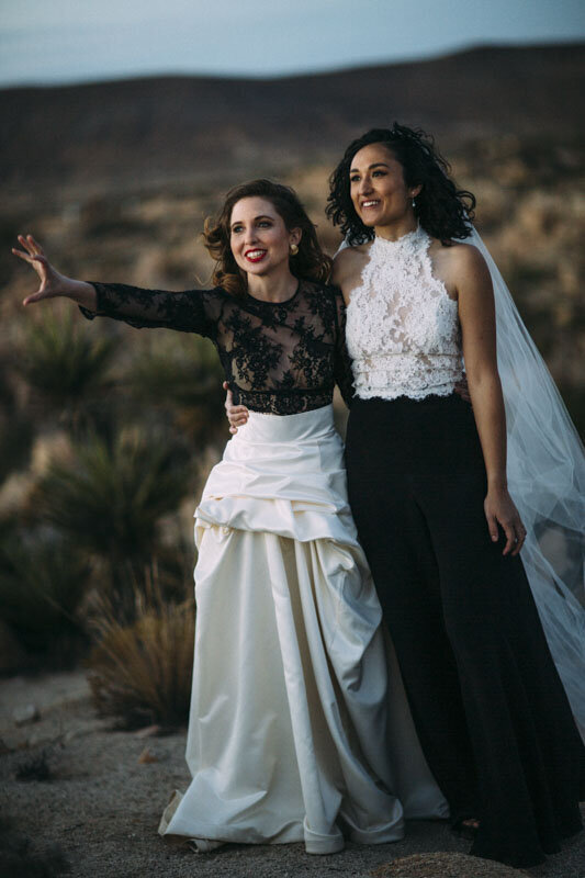 Two brides embrace in the desrt on their wedding day. They are wearing black and white two piece bridal fashion.