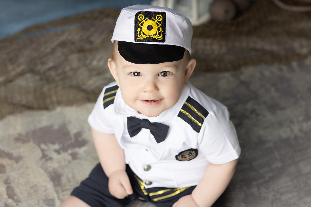 Close-up of a baby boy in a white and navy sailor outfit with a captain’s hat, looking up and smiling during his nautical-themed milestone session, captured by a Dallas milestone photographer.