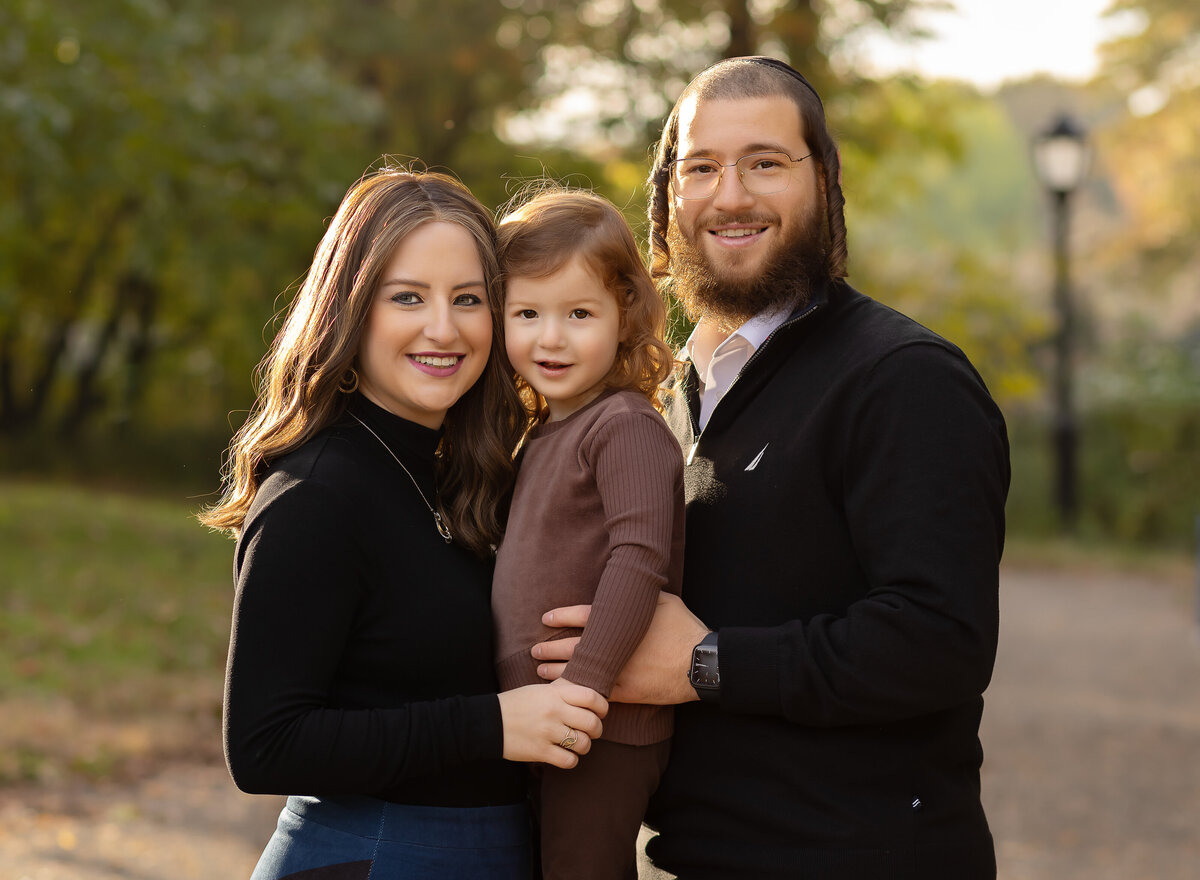 Mom, dad, and toddler are captured in a brooklyn ny park. Mom is holding the little boy and the little boy is between the mom and dad. All are smiling at the camera. Captured by brooklyn family photographer Chaya Bornstein.