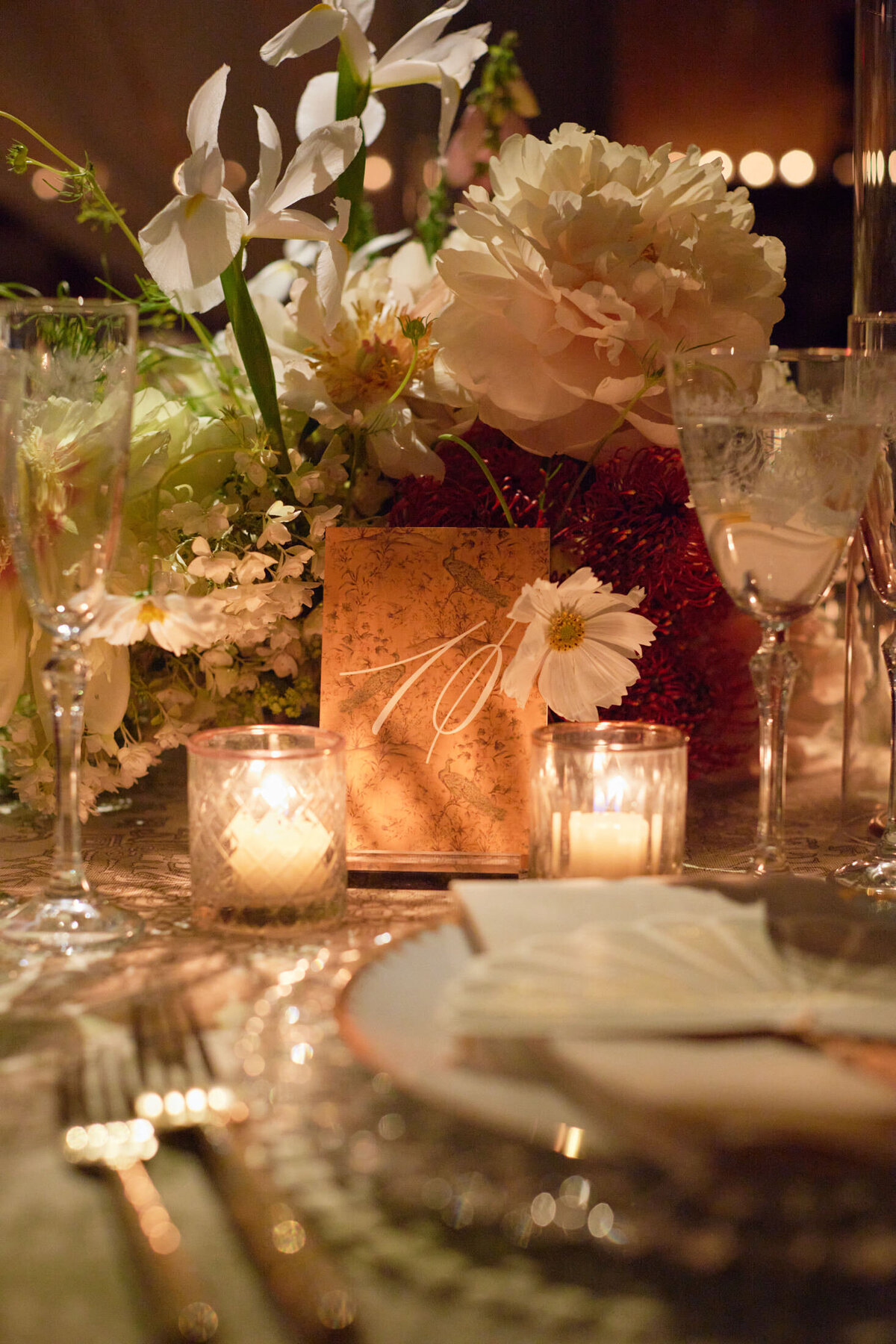 The photograph captures a beautifully set table with an elegant floral centerpiece, featuring white lilies and peonies. The setting exudes a warm and romantic ambiance, complemented by the soft glow of lit candles in glass holders. Nestled neatly alongside the folded napkins on a plate is a decorative menu card, creating a captivating scene at the New York Public Library, as captured by photographer Shawn Connell. Videographers for the event were provided by Aaron Navak Films.
