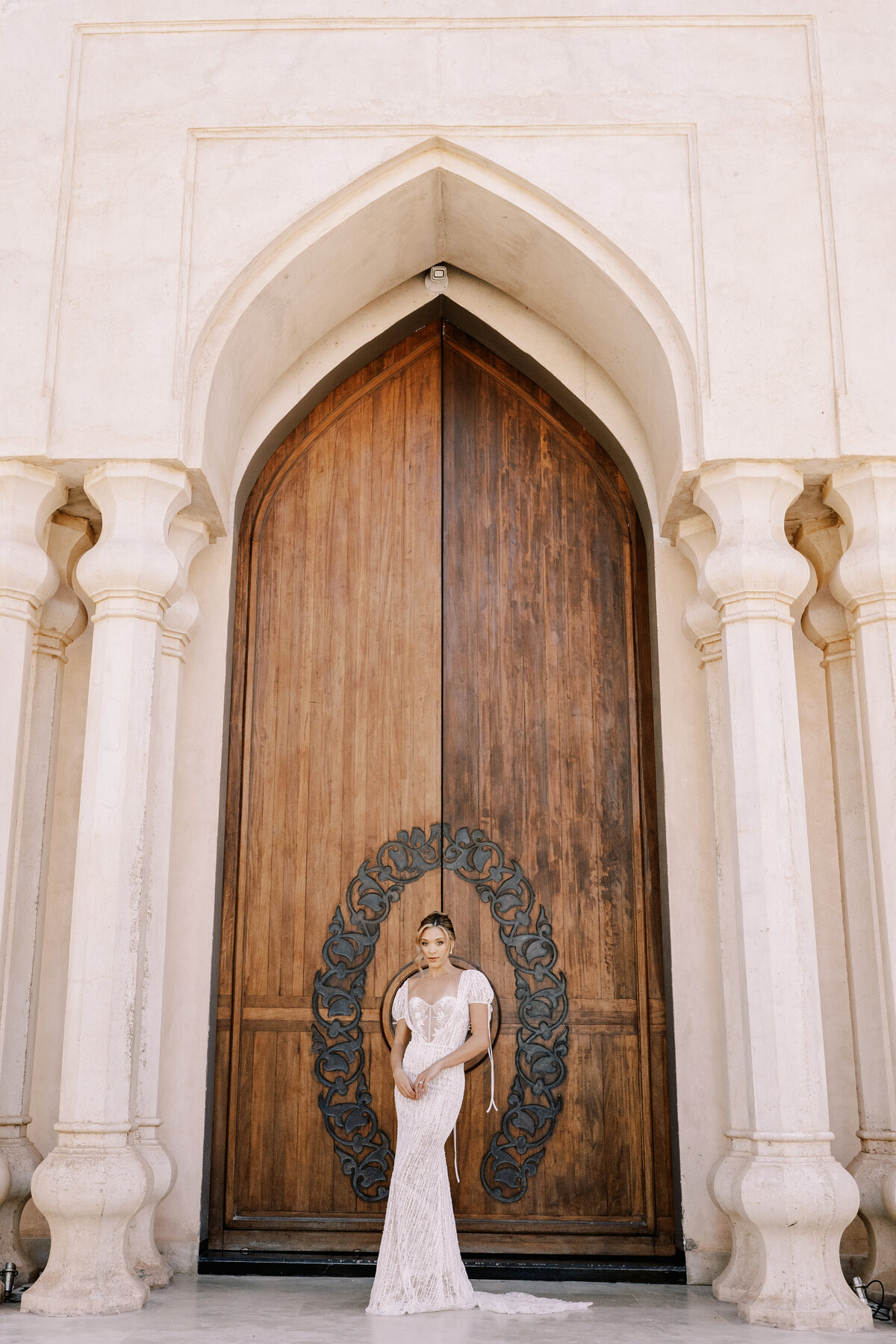 Bride in updo and short sleeved beaded wedding gown with bows in front of large wooden embellished doors at Palais Namaskar Morocco for a destination wedding photographed by Italy photographer