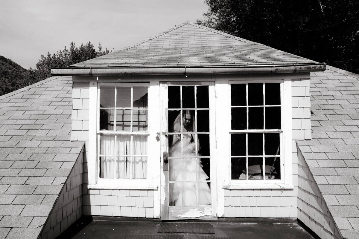 Black and white photo of the bride inside a room, with a french door, from the outside at The Ausable Club, New York.