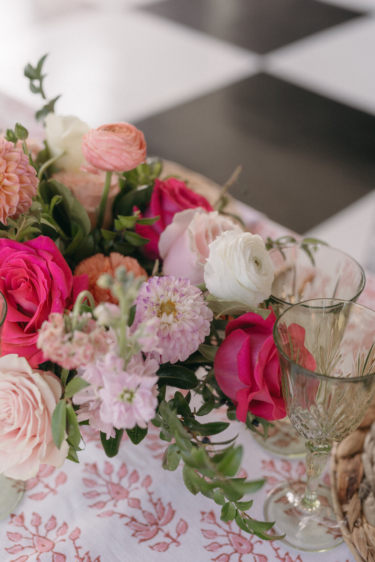 Wedding reception table centerpiece featuring garden roses, dahlias, and ranunculus in bright pink and peach tones paired with vintage glassware and patterned linens.

