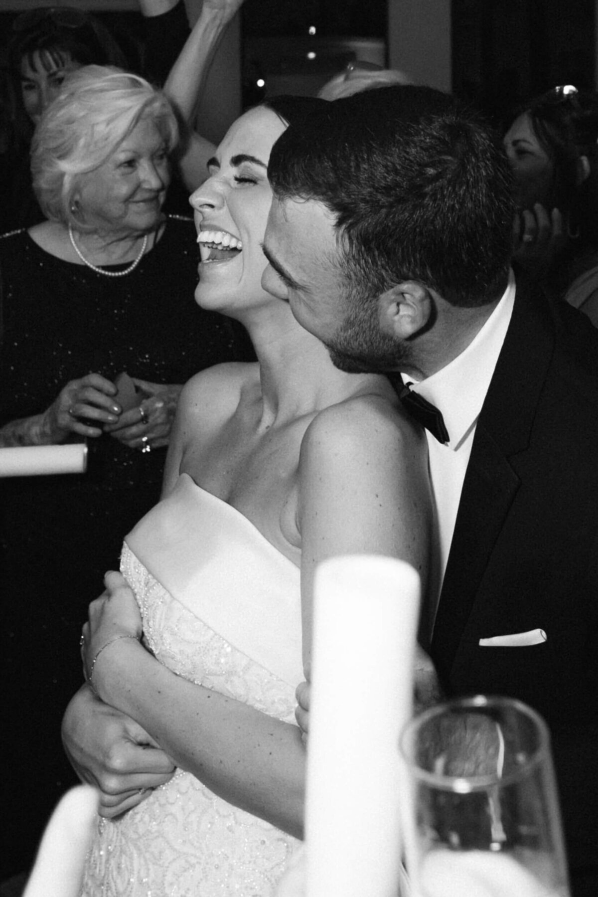 A candid black and white  wedding photo of the groom kissing the bride while she smiles and laughs