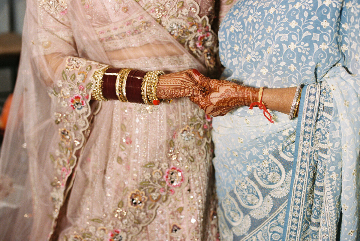 Bride in embroidered lehenga holding hands with family member in blue sari, showcasing wedding henna and bangles.