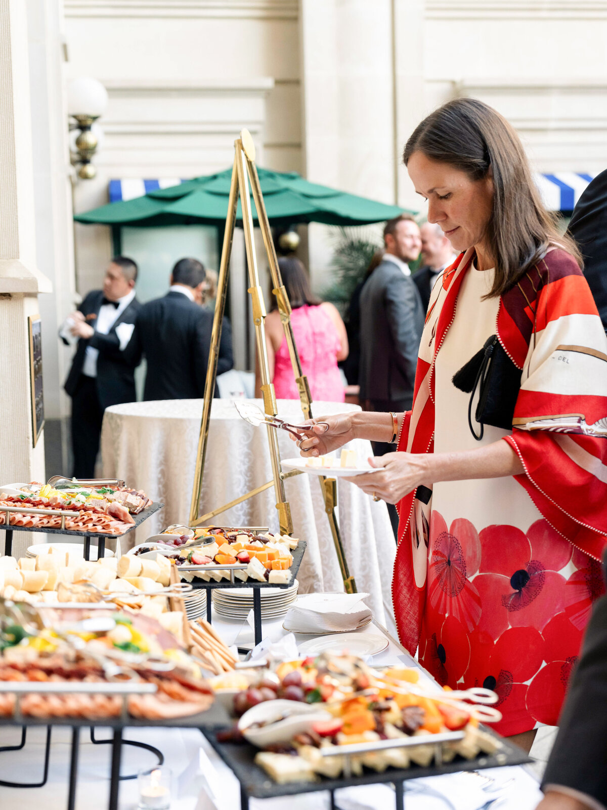 A woman in a floral dress serves herself food from a buffet table at an elegant outdoor event. The table is filled with various appetizers, and other attendees are seen in the background near a green parasol.