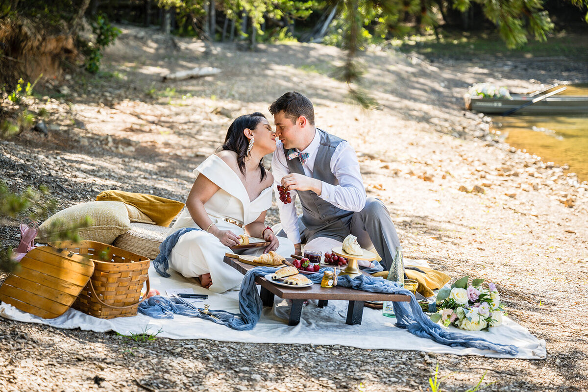 A couple sits down for a brunch picnic on their elopement day along the shores of Carvin’s Cove in Roanoke, Virginia. The picnic setup includes a picnic basket, a couple of pillows, a small table with a draping blue fabric, and all of their food, including croissants, various fruits, cakes, jam, and honey. Each person in the pair holds onto a piece of food as they rub noses.