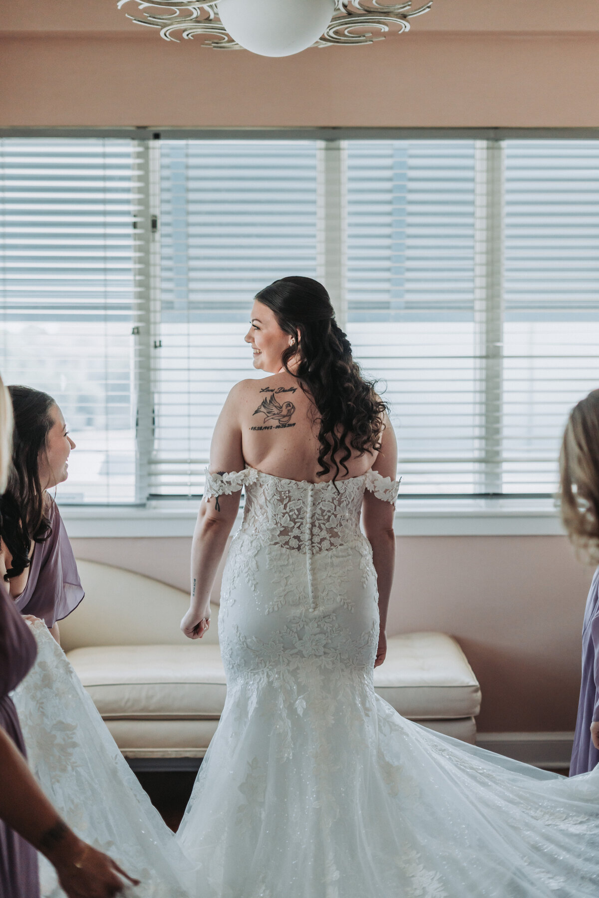 A bride is seen from behind in her stunning off-the-shoulder lace wedding dress, looking over her shoulder with a smile, showcasing the intricate details of her gown and back tattoo, in a bright room at the Lesner Inn in Virginia Beach, captured by Quinn Photography LLC.