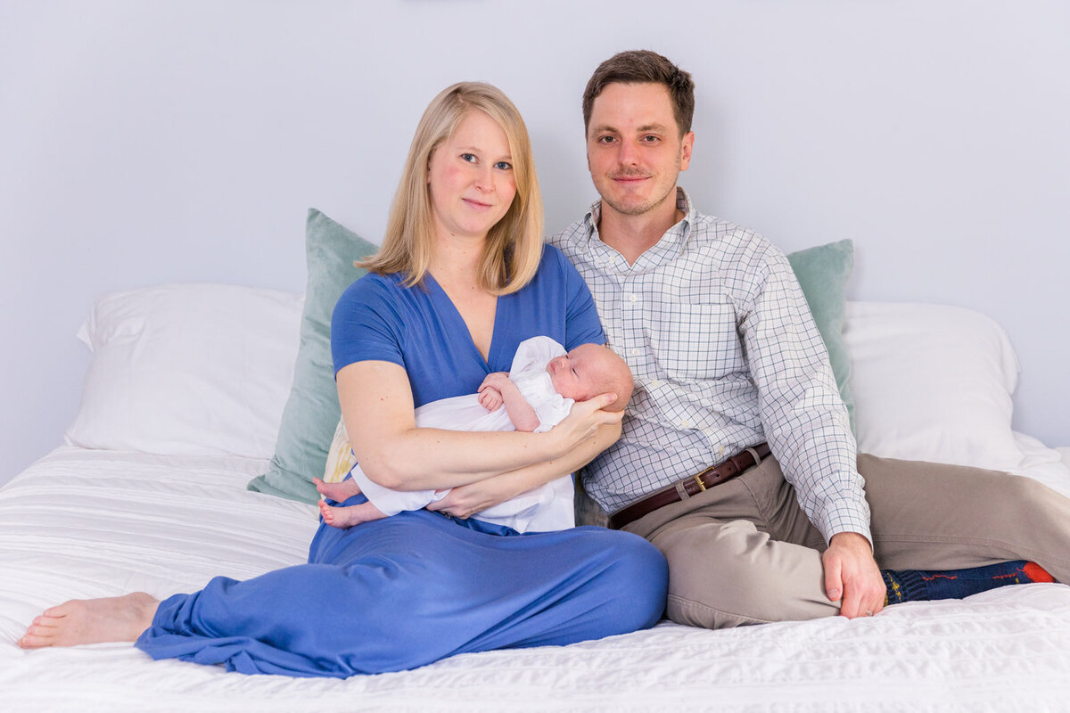 parents holding newborn baby girl sitting on a bed mom wearing a long blue dress Laure Photography Atlanta family and newborn photographer