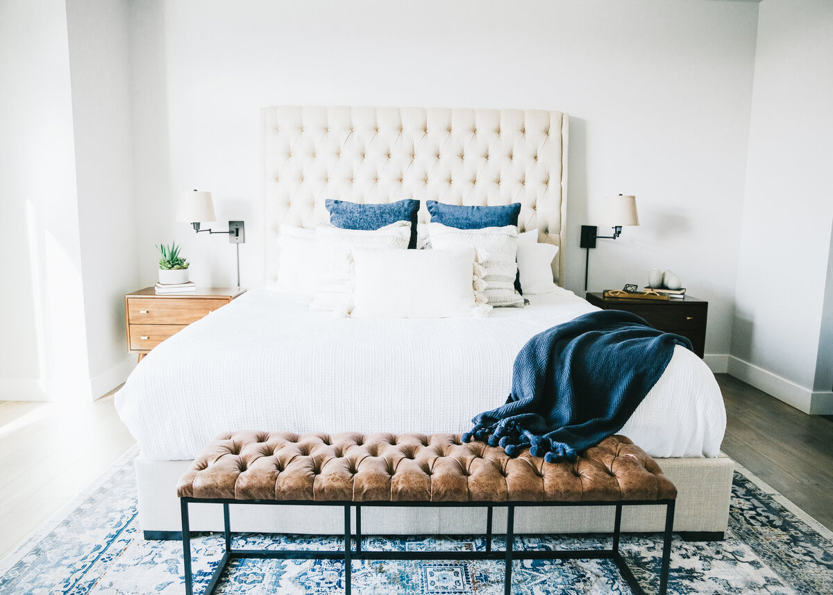 A white painted bedroom features a padded headboard white sheets, with a navy throw and a tan leather bench at the end.