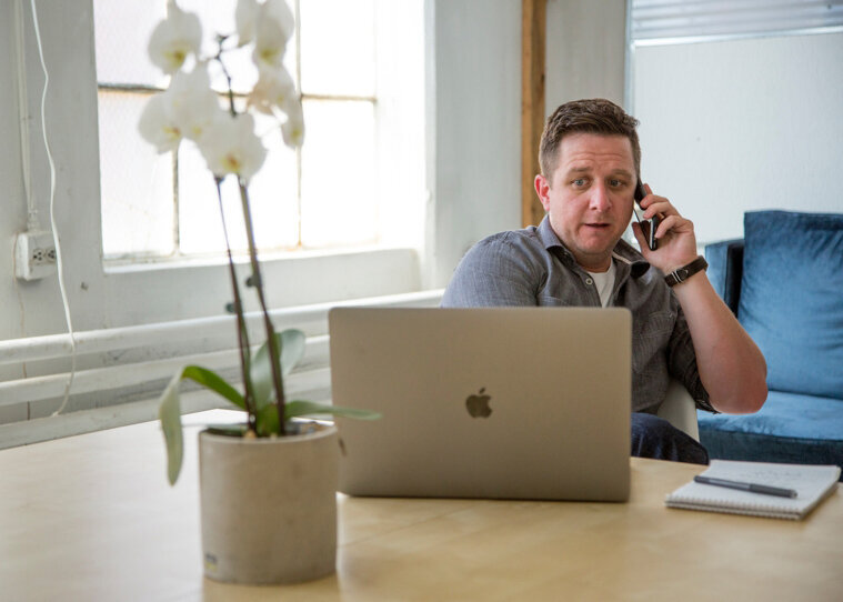 A male business owner sitting at his desk, looking at his laptop while on a phone call, and posing for a South Shore MA branding photographer 