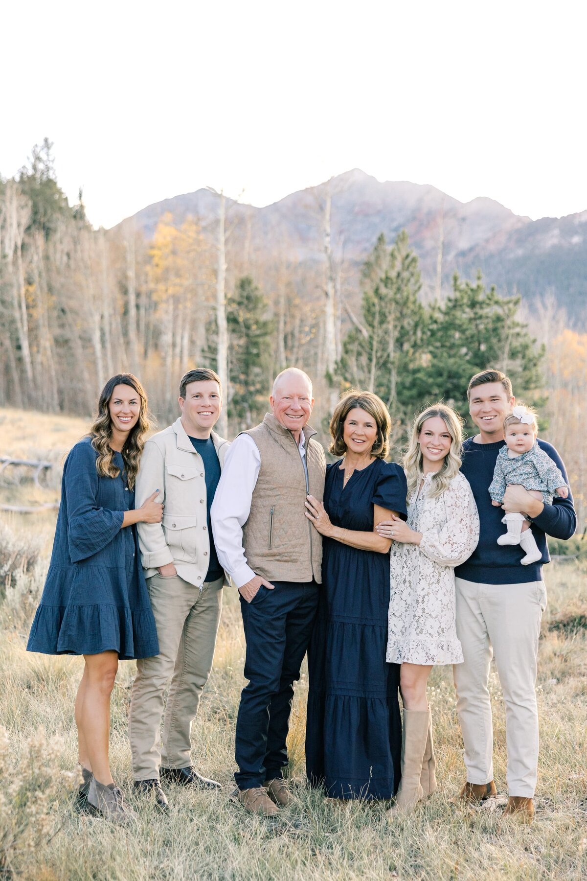This photo shows the whole extended family with Grandma, Grandpa, the family of three with their baby and a son with his girlfriend. There are tall mountain peaks behind them. 