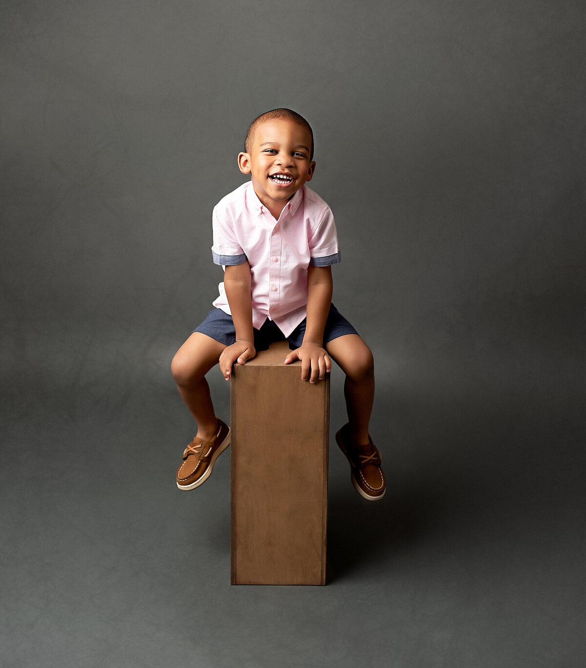 A little boy sitting on  an apple box with hands on the box with one leg on each side.  The boy looking at the camera smiling.