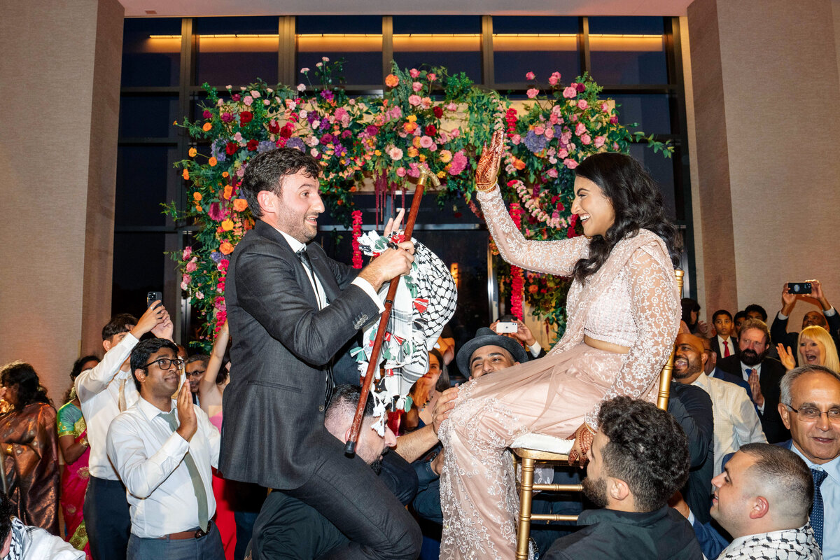 A joyful couple is lifted on chairs during a celebratory event. The man holds a decorated cloth, and the woman waves. They are surrounded by cheering guests, with vibrant flower decorations in the background.