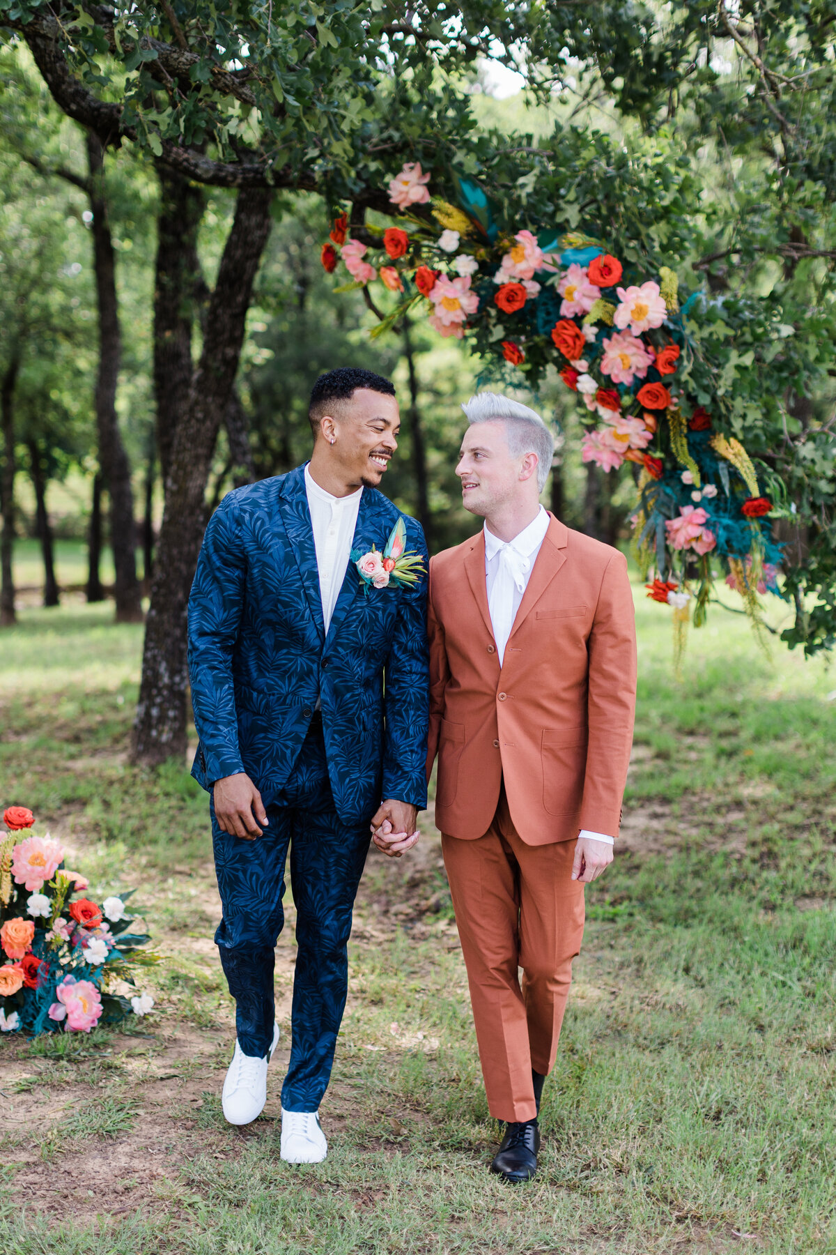 Portrait of two grooms walking through the woods on their wedding day in Dallas, Texas. The groom on the right is wearing a dark orange suit while the groom on the left is wearing an intricately patterned blue floral suit with a boutonniere. Above and to the side of them are large, colorful floral arrangements.