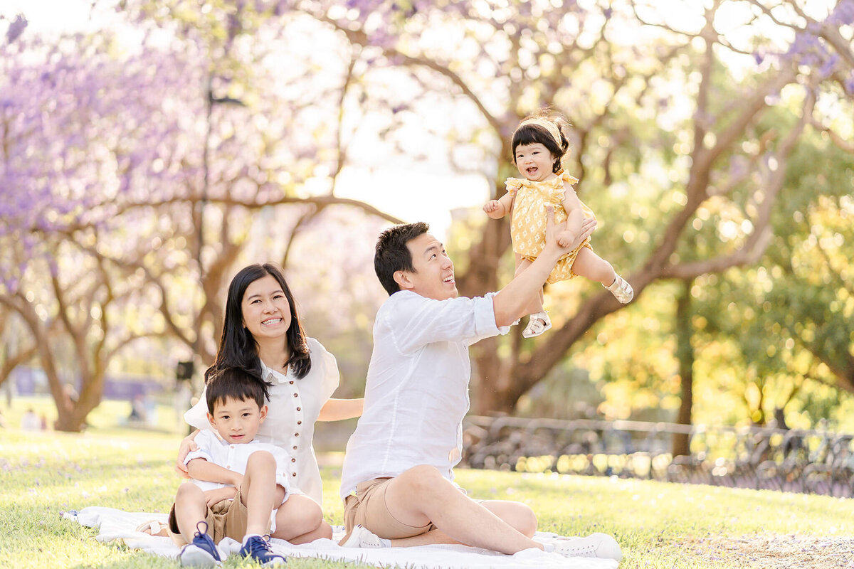 family relaxed on picnic mat under jacarandas in brisbane hotspot flying baby in air
