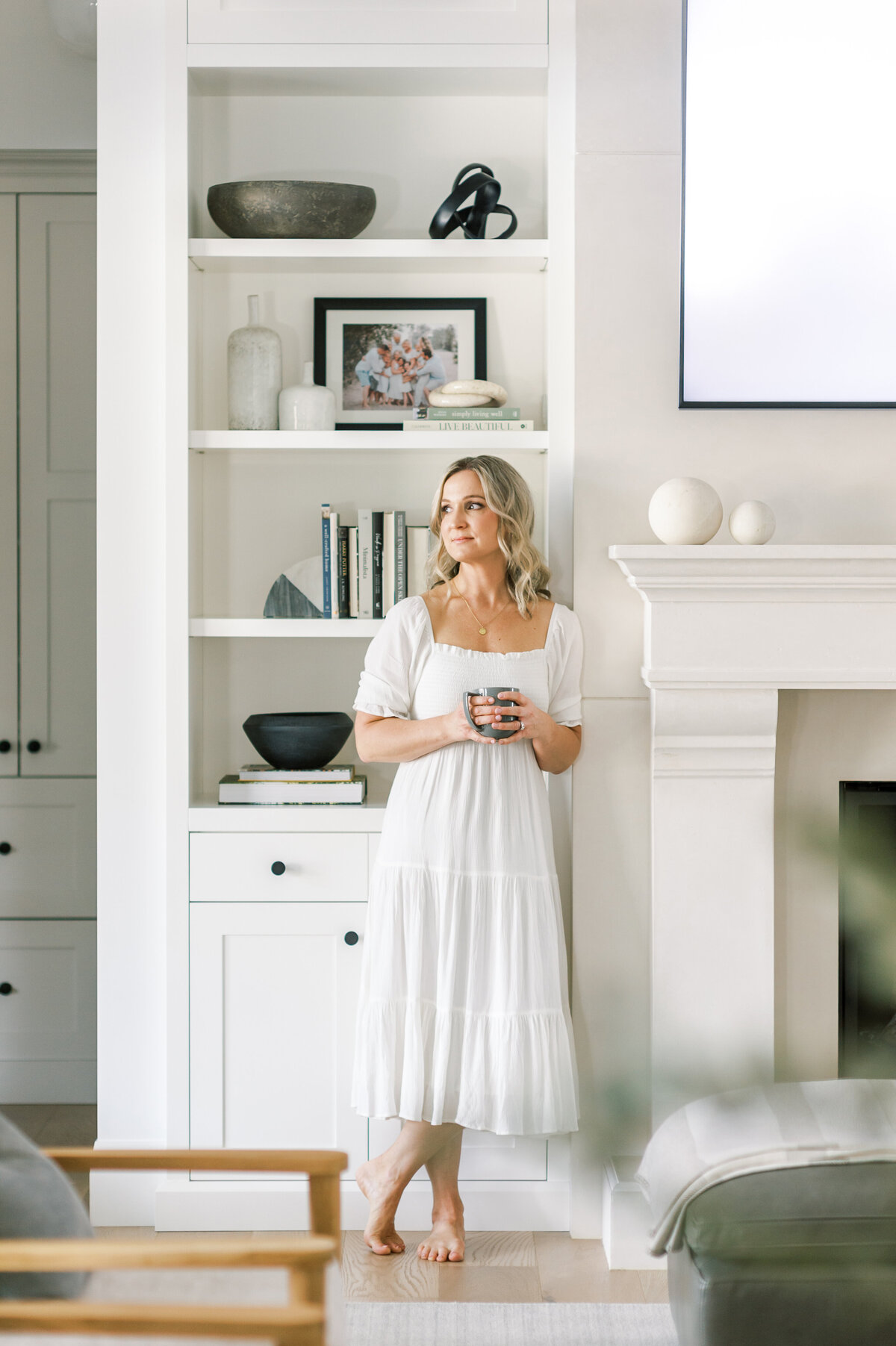 Premiere London, ON interior designer is wearing a long white dress and leaning against her oversized white fireplace mantle. She is looking over her shoulder toward  the cascading windows with styled bookshelves behind her.