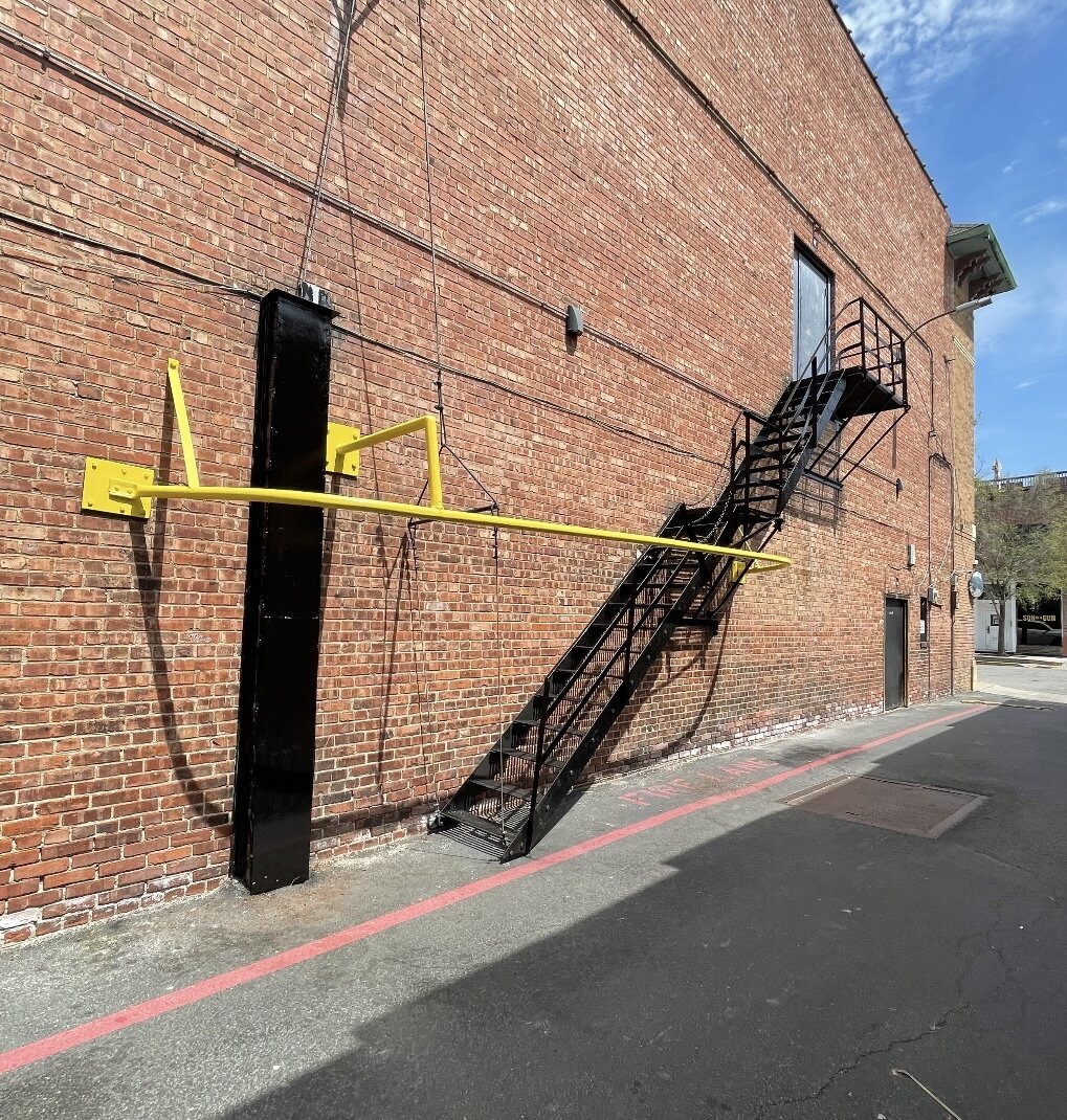 Black steel fire escape staircase with welded yellow safety rail installed on a brick commercial building exterior.