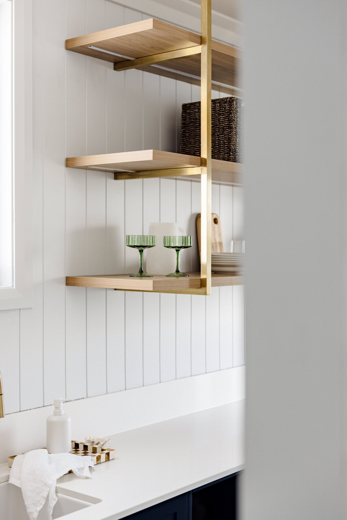 Renowned Newcastle Interior Designer brings a minimalist touch to this kitchen, featuring wooden shelves, green glasses, and a woven basket on a paneled white wall.