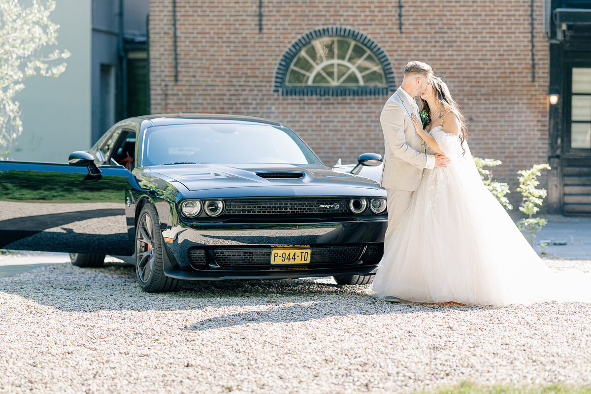 Standing next to a Dodge charger, a bride and groom kiss during their portraits after the ceremony near Rotterdam