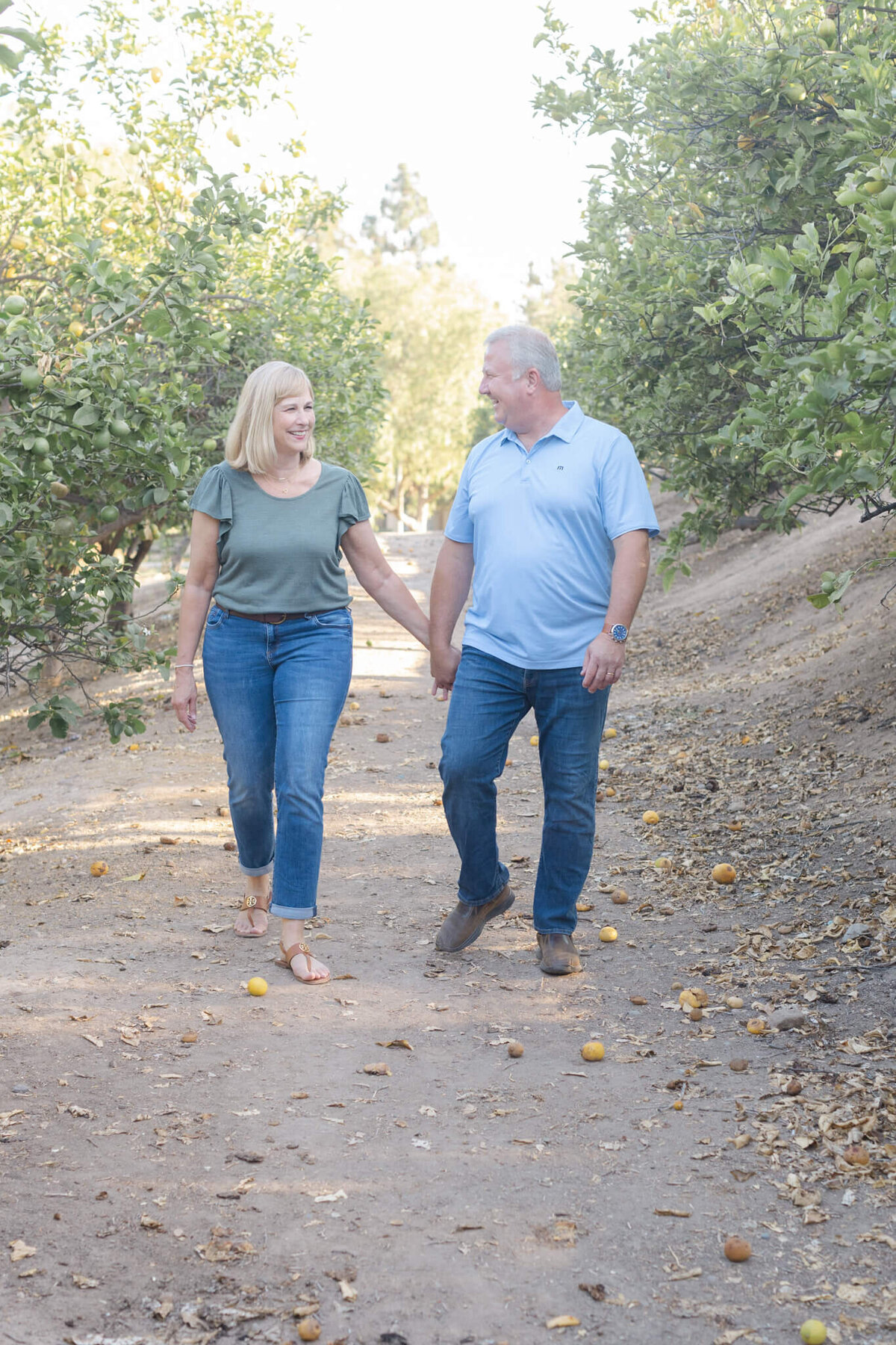 A woman with blonde hair wearing a green shirt and blue jeans looking at her husband and holding his hand as he's wearing a light blue collared shirt and blue jeans, looking at her and walking towards the camera between two groves of lemon trees in Tustin.