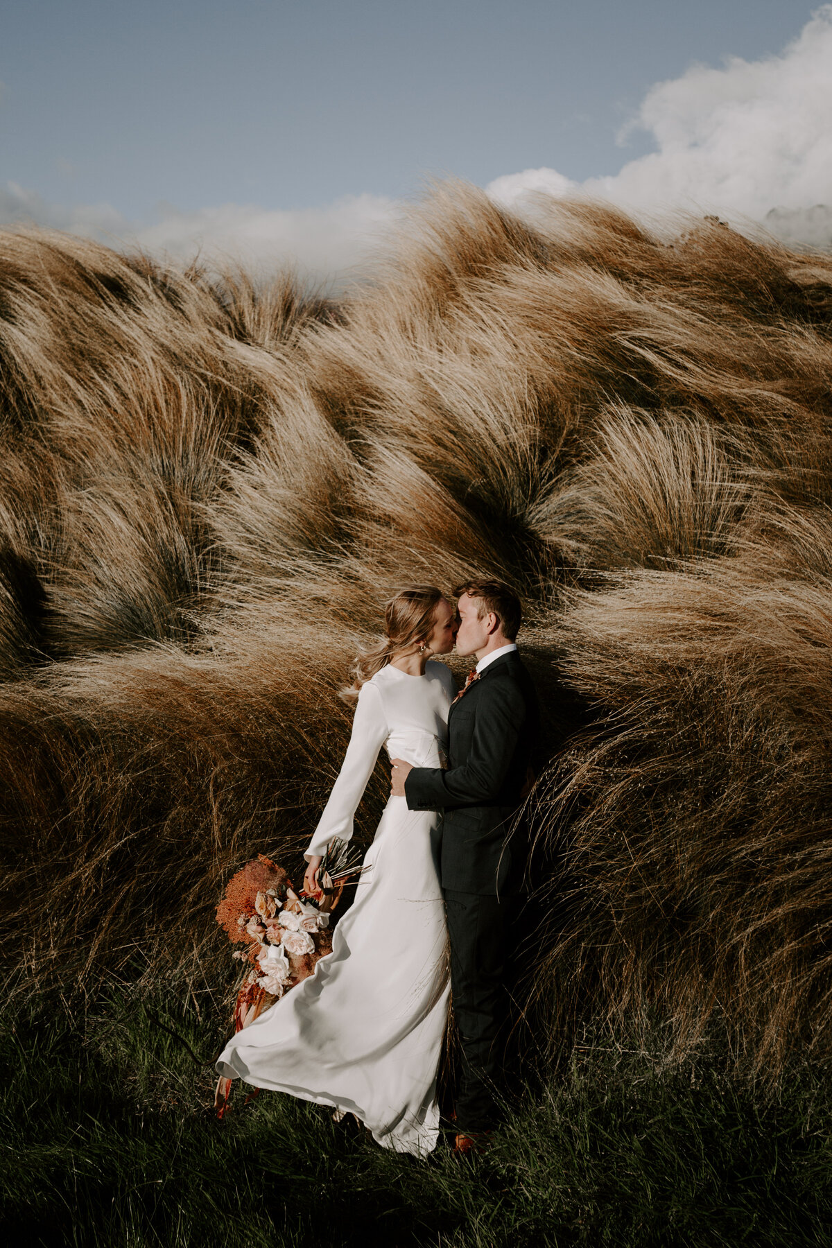 The Lovers Elopement Co - bride and groom kiss surrounded by long grass - Jack's Point wedding Queenstown venue
