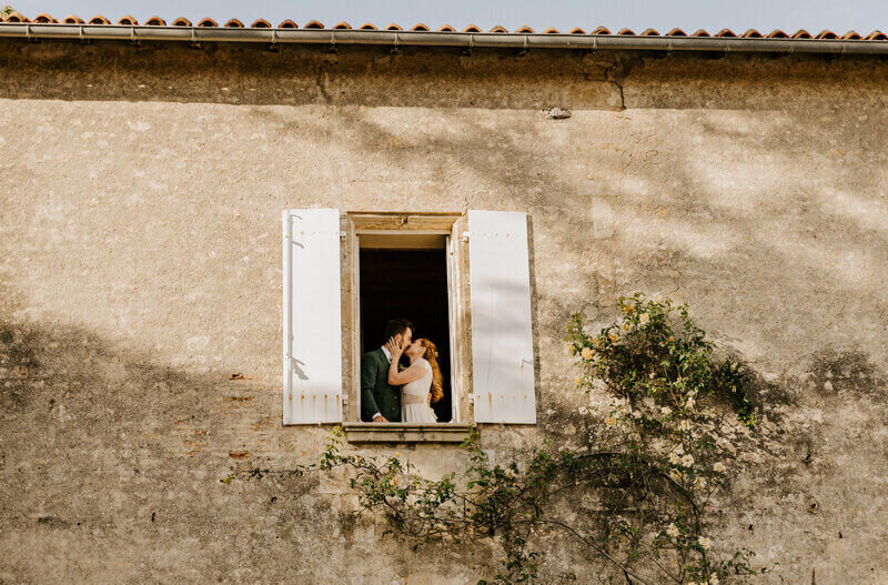 Mariés s'embrassant au loin dans l'encadrure d'une fenêtre aux volets en bois blanc d'une bâtisse en pierre pour une shooting photo mariage en vendée.