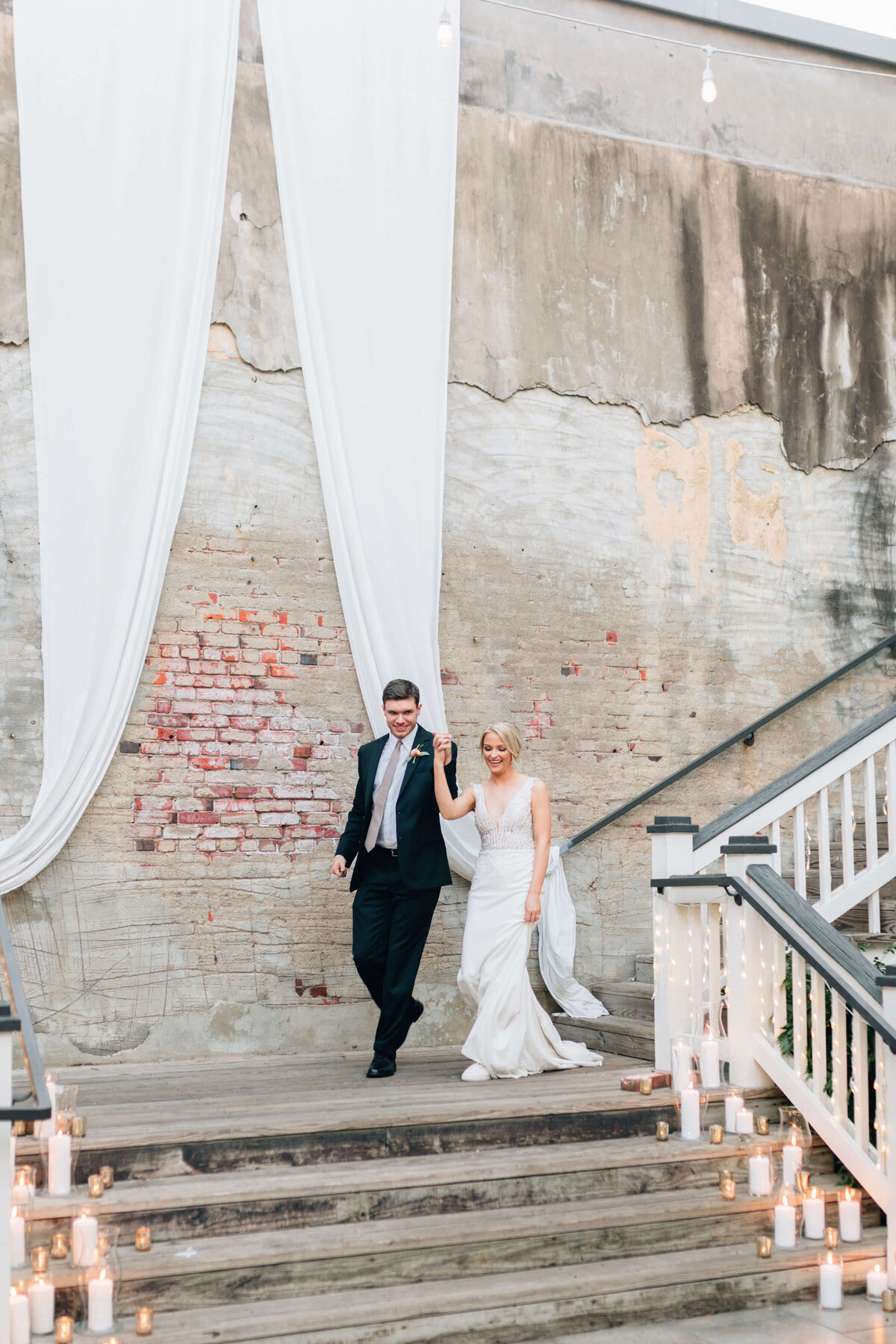 A bride and groom hold hands while walking down outdoor steps decorated with candles. They are smiling, beneath white drapes and in front of a textured brick and concrete wall.