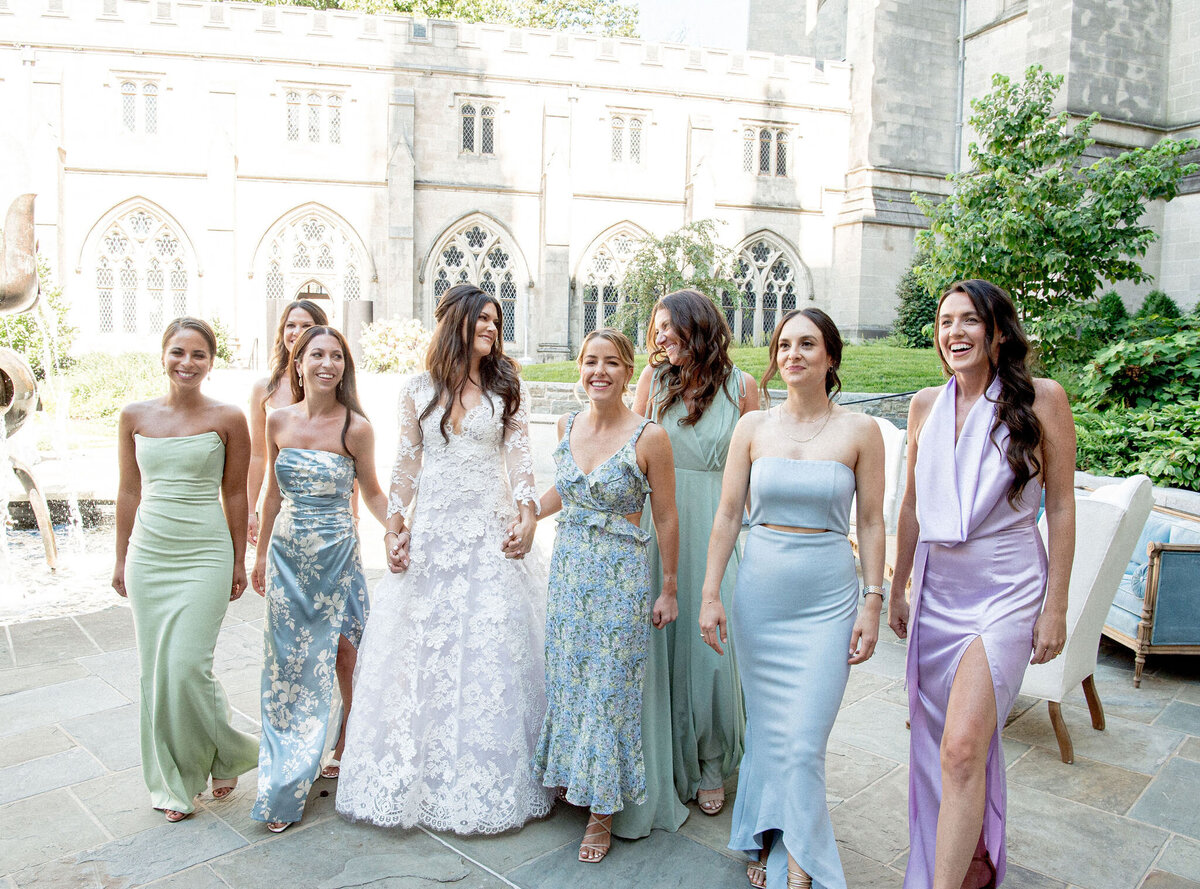 The photo was taken at Washington D.C., Washington National Cathedral by photographer Twah Dougherty. The bride in a white lace wedding dress walks hand-in-hand with her bridesmaids, who are wearing different shades of pastel-colored dresses, outside in a courtyard with stone architecture in the background. The group is smiling and appears to be in a celebratory mood, capturing a beautiful moment against the stunning backdrop of the cathedral. Additionally, Current Films served as the videographers for this special occasion, capturing the joyous moments on film.