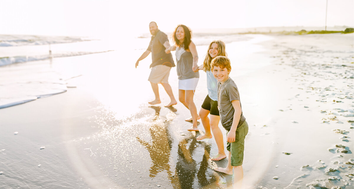 A family smiles at camera while holding each other's hands and running towards the ocean waves with the sunset behind them.  By SAVI  Photography - San Diego California Photographer