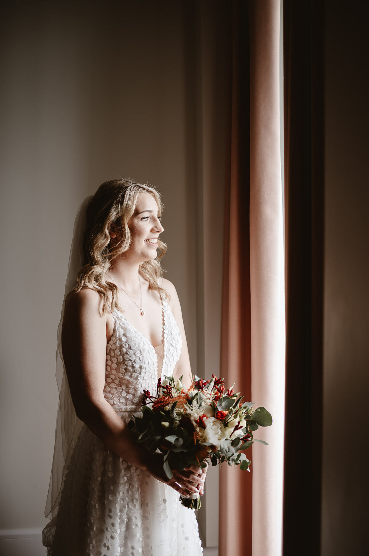 bride smiling and holding flowers whilst looking out the window