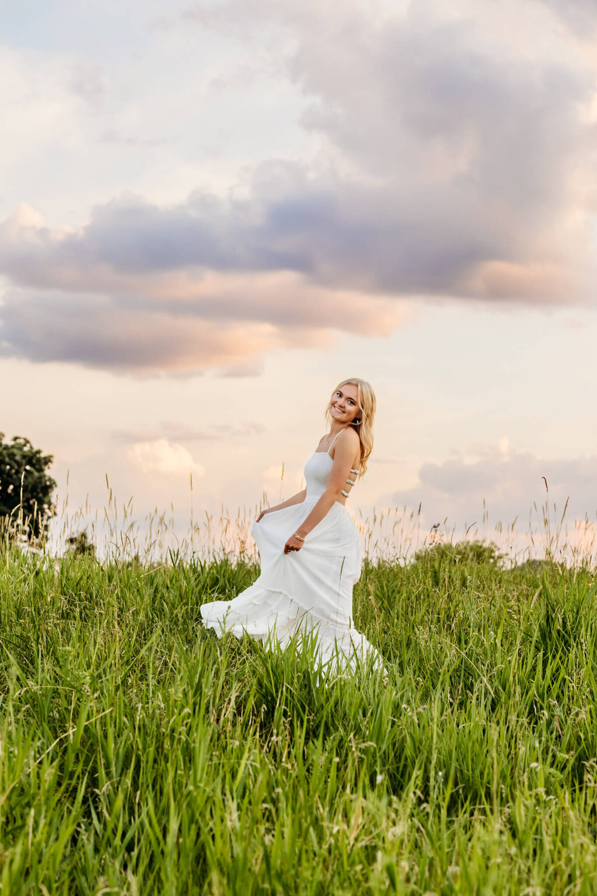 blonde high school senior girl in a white flowy dress spinning on top of a hill with cotton candy colored clouds behind her captured by Ashley Kalbus Photography