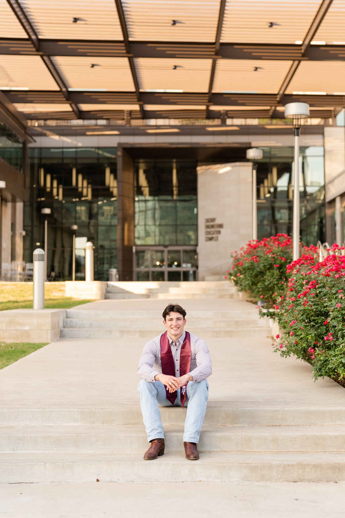 Texas A&M senior guy sitting on step and leaning on knees while wearing white wash jeans and maroon button down with maroon stole in front of Zachary Engineering Complex