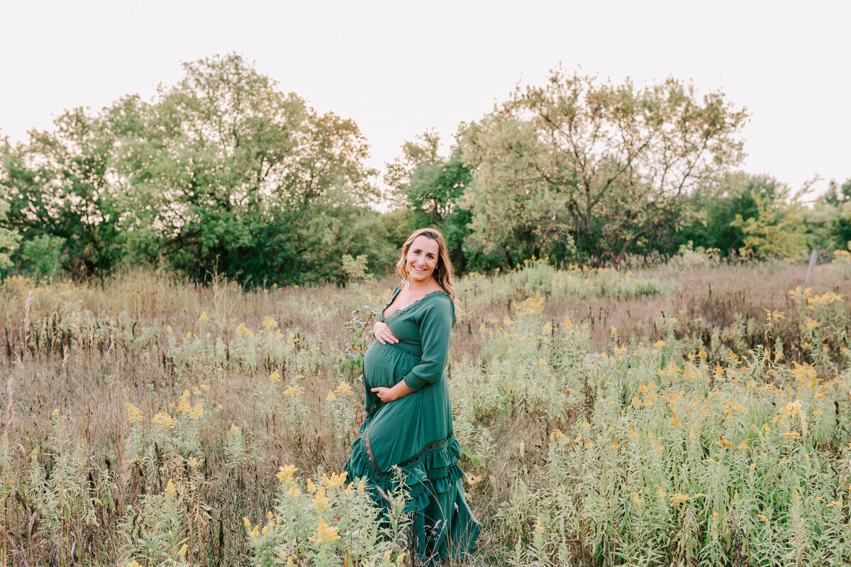 Mom smiles while holding her belly in a field during her session with Chelsey Kae Photography.