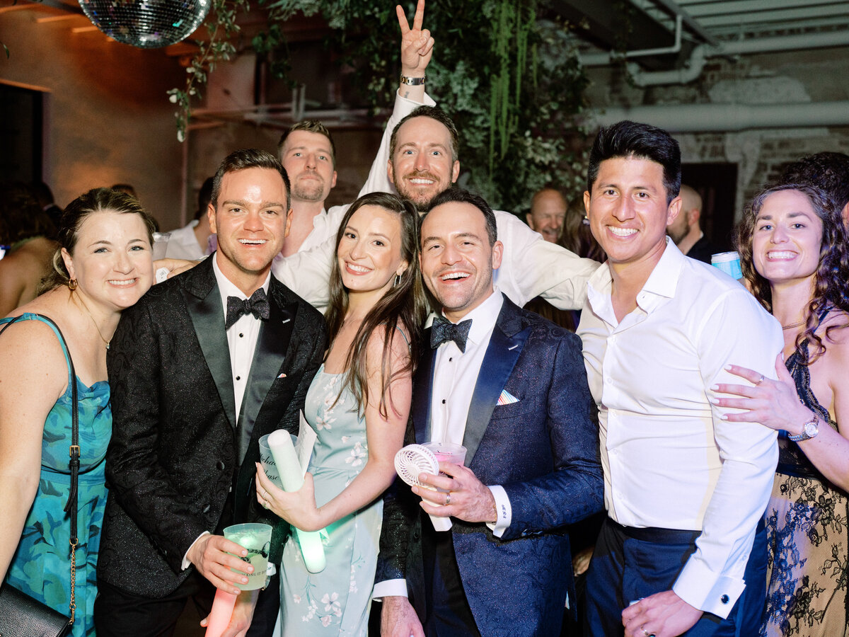 A lively group of friends in formal attire dance joyfully, holding colorful drinks and illuminated props, celebrating together at a wedding reception filled with excitement.