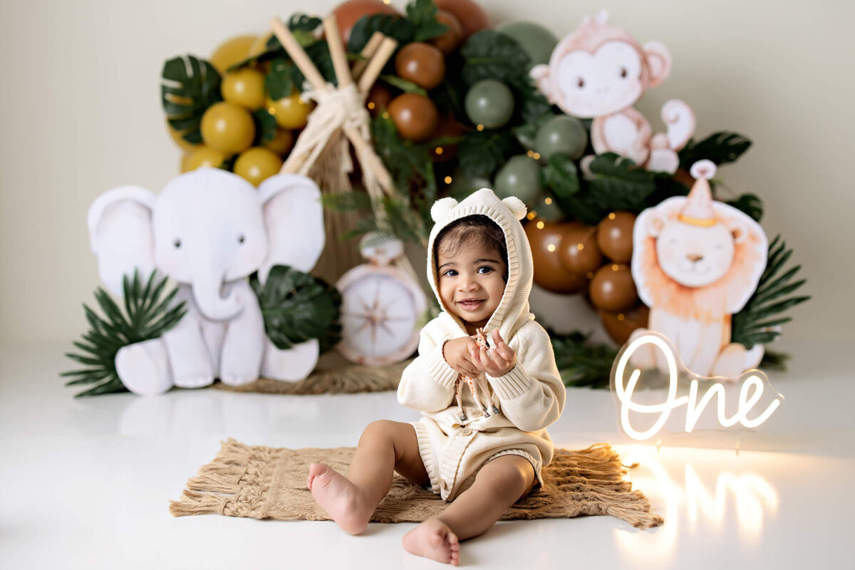 A smiling toddler in a cream-colored hoodie with bear ears sits on a mat. Captured by a Bloomington Illinois newborn photographer, jungle-themed decorations and a glowing "One" sign create the perfect backdrop for this adorable milestone.