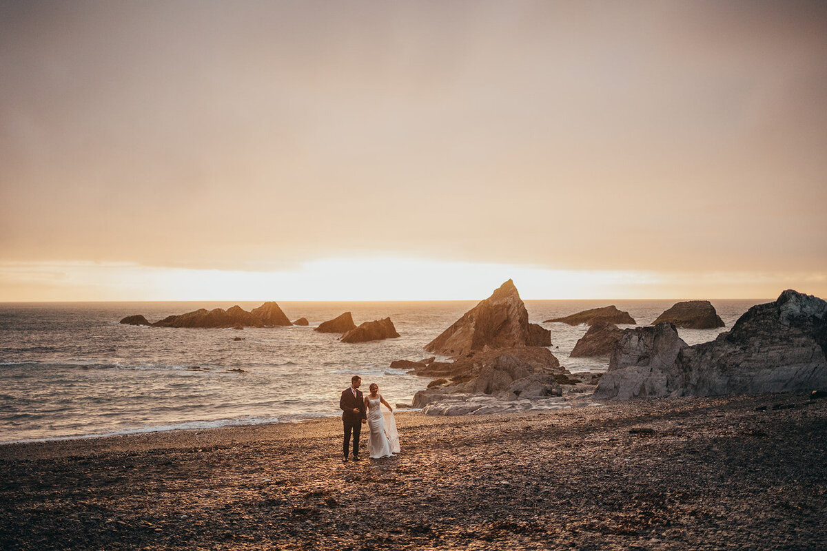 Bride and groom on a grey slate beach at sunset with craggy rocks coming out of the sea.