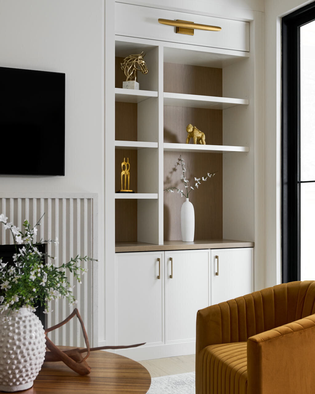 A stylish living room featuring custom built-in cabinetry by Barzotti Woodworking in Oakville. The white shelving unit with light wood backing is accented by gold decor, integrated lighting, and modern hardware, complementing the elegant interior design.