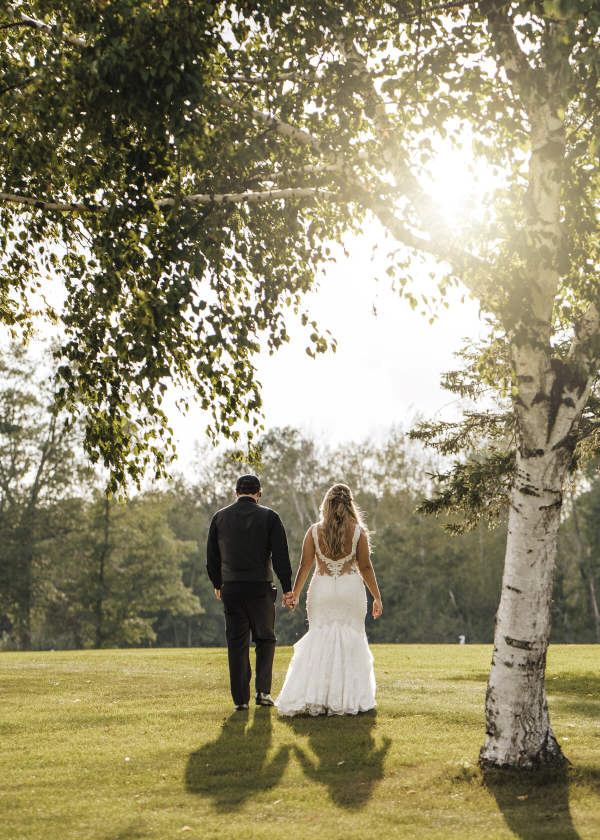 A couple holding hands, walking through a sunlit golf course, with the bride in a white dress and the groom in a dark suit, signifying a moment of togetherness and new beginnings taken by jen Jarmuzek photography a Minneapolis wedding photographer