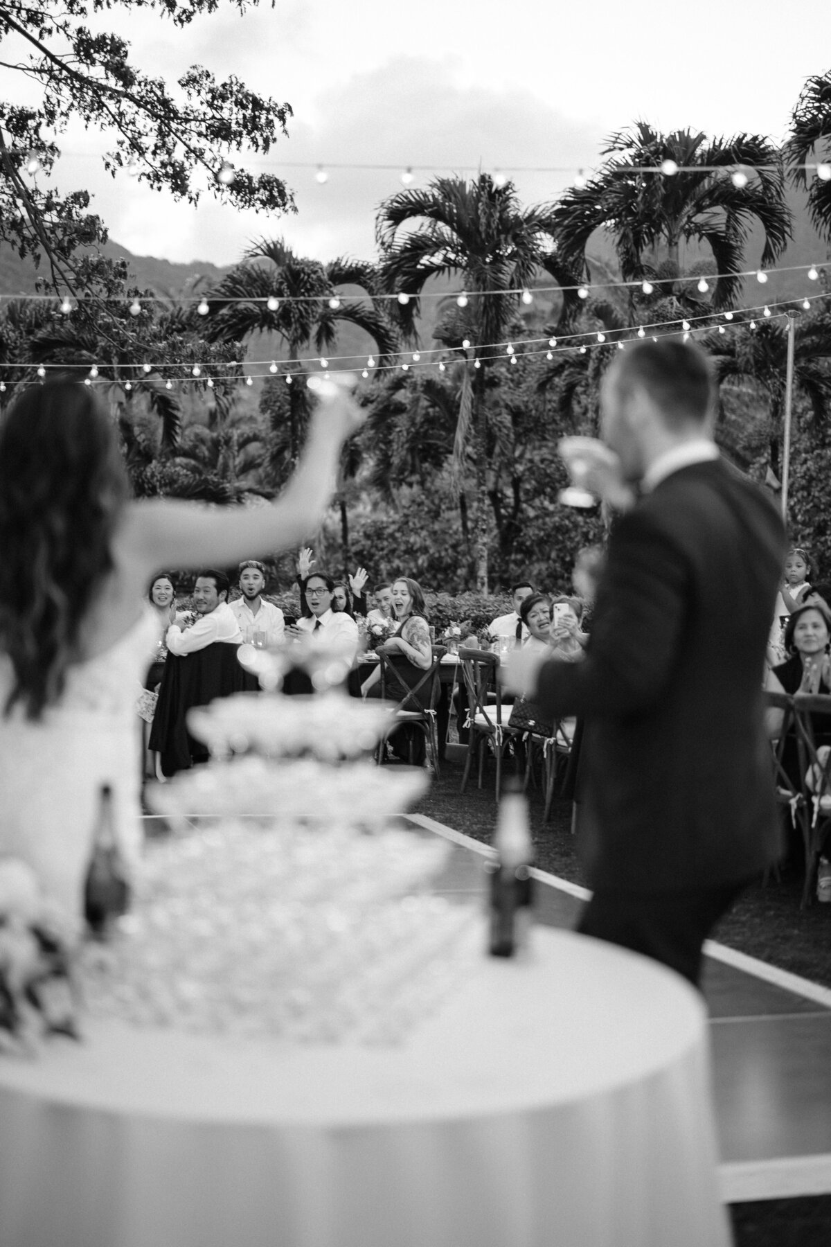 Bride and groom cheering in front of an out of focus champagne tower to their guests in black and white