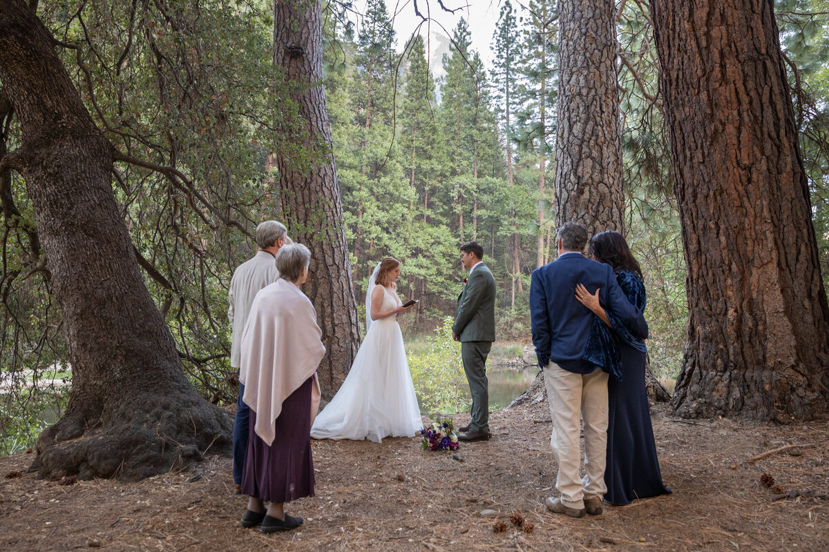 A group of people stand in a semi circle while the bride reads her vows to her groom on their elopement day.