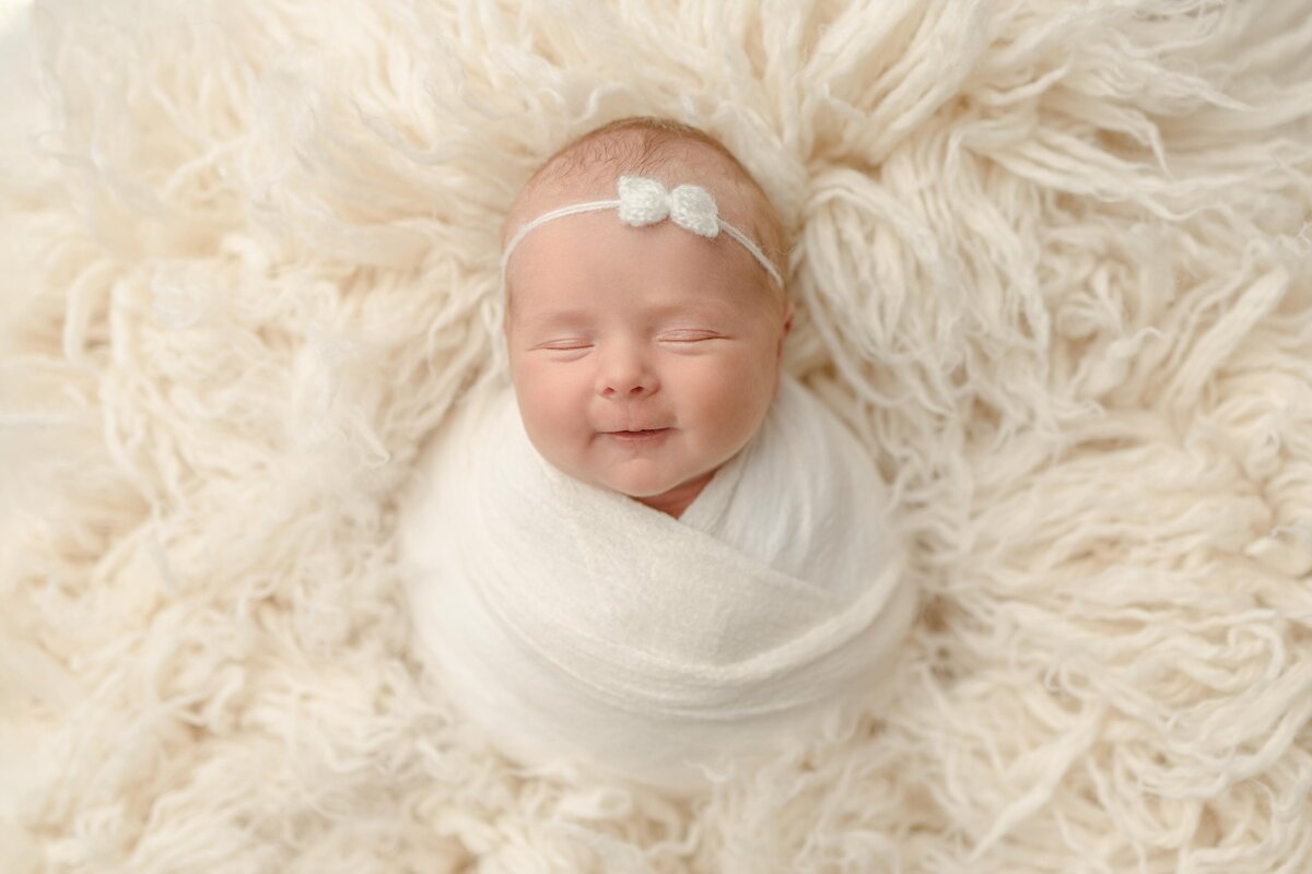 Newborn baby swaddled in white laying on white flokati smiling