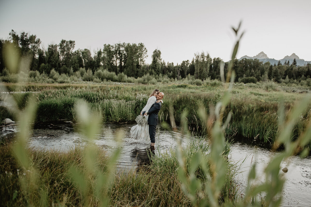 jackson wyoming photographer captures wyoming elopement in the grand tetons with groom picking up his bride while they walk together through the snake river