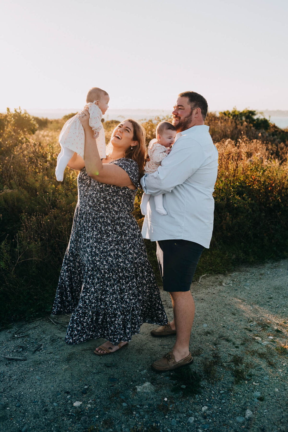 A mother holds up her baby as her husband, who is holding their other twin, looks on and smiles during their photoshoot with Boston family photographer, Allison Wolf