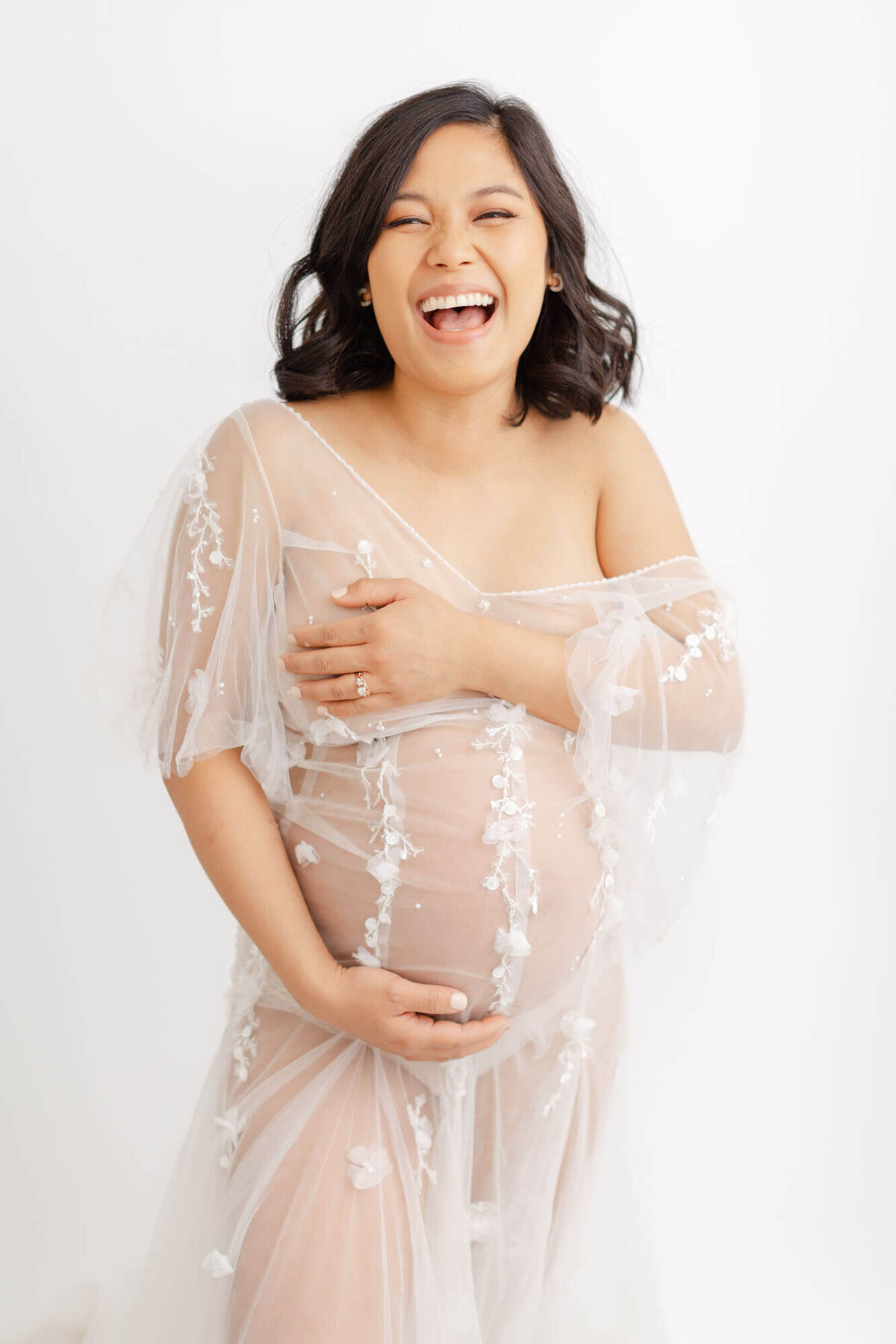 Pregnant Woman laughing big towards the camera. She has dark shoulder-length hair and is wearing a see-through tulle dress with some hand beaded work. She is holding her chest with one arm and holding her belly with the other hand.