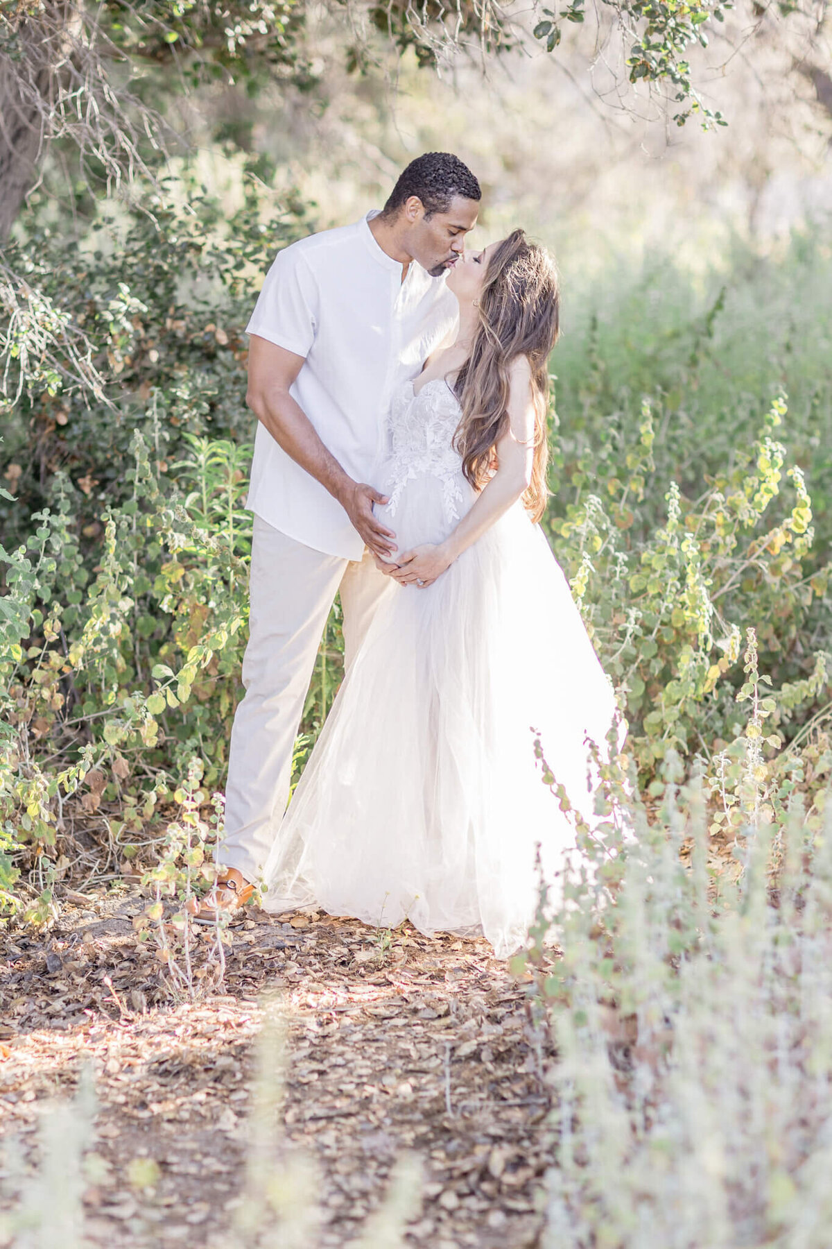 A couple kisses in a sunlit forest, surrounded by green foliage. The woman wears a white dress, and the man wears a white shirt and beige pants. They stand on a path amidst the soft, dappled light filtering through the trees.
