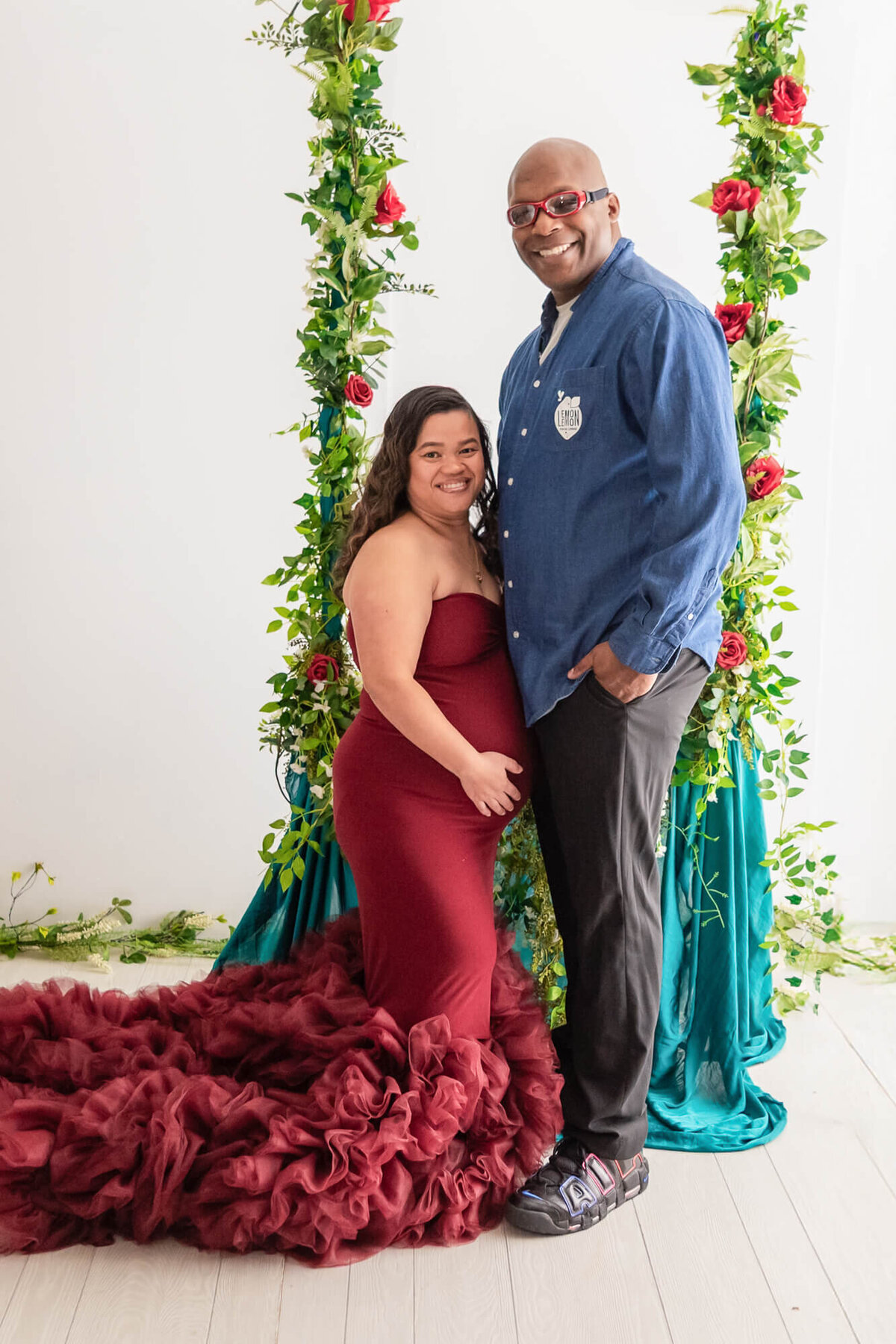 A happy couple stands together in front of a vibrant floral backdrop, celebrating their pregnancy with joy and love in Youngstown photography studio