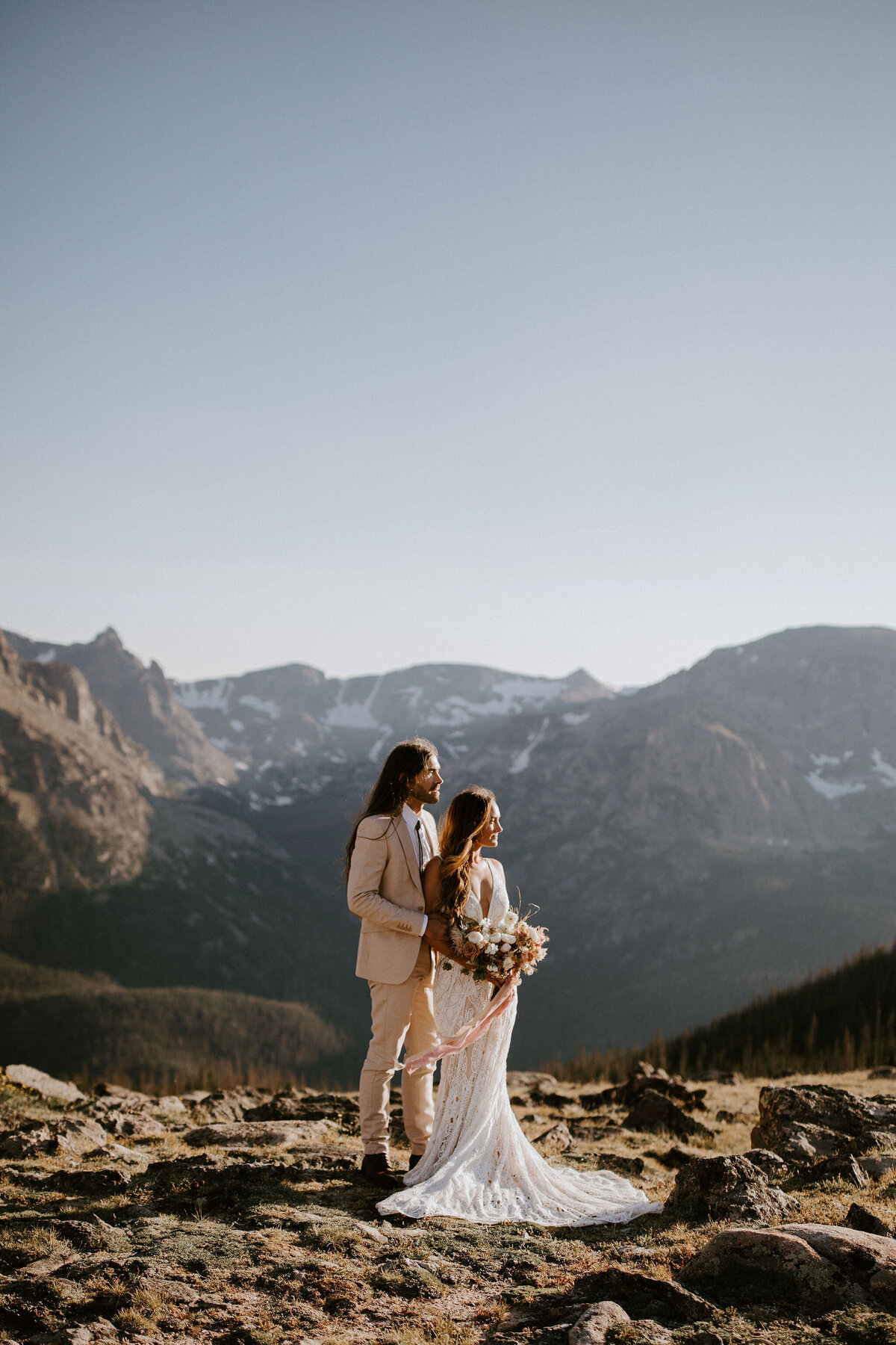 Side profile of standing Bride and groom wearing an ivory suit and white wedding gown with mountains in the background.